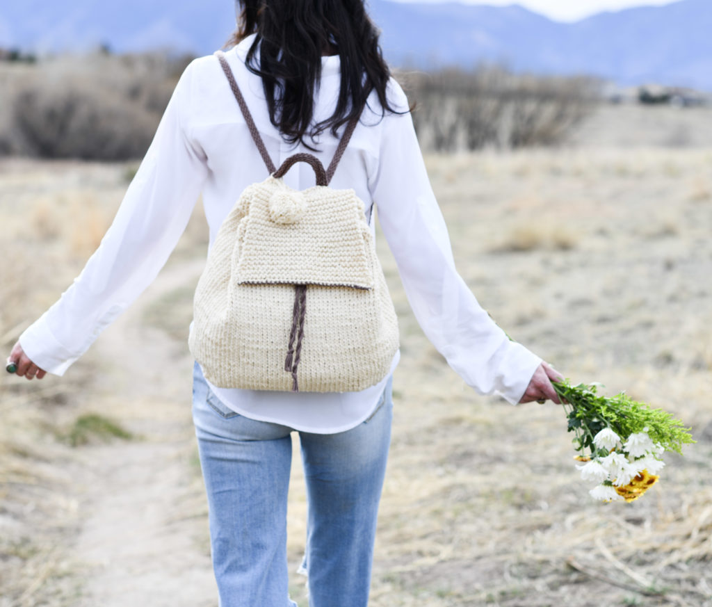 a women wearing a knitted bag on her back, holding wildflowers in one hand. 